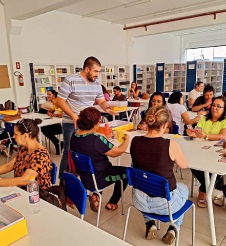 MEC: Grupo de professores participa de formação docente em mesas dentro de uma biblioteca, manuseando cartas e materiais educativos.